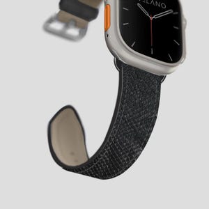 May include: A silver smartwatch with a black screen and a black textured leather strap. The watch has an orange accent on the side. The strap is curved, showing the light tan underside.