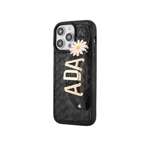 May include: Black phone case with a textured, scale-like pattern. The case features a gold-coloured daisy and the name "ADA" in gold letters. The case has a strap for holding the phone.