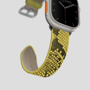 May include: A silver smartwatch with a black screen and a yellow and black snakeskin-patterned strap. The strap is made of a flexible material and has a yellow underside. The watch has an orange button on the side.
