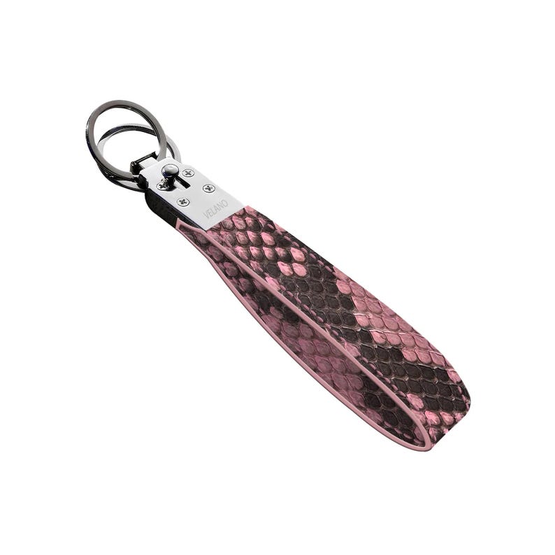 May include: A pink and black snakeskin-patterned keychain wrist strap with a silver-tone metal clasp and key ring. The strap is made of a textured material and features the brand name "VELANO".