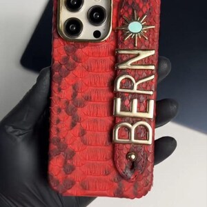 Może przedstawiać: Czerwone etui na telefon o fakturze skóry węża ze złotymi akcentami. Posiada złoty wzór promieni słonecznych z turkusowym środkiem i napisem "BERN" w złotych literach. Etui ma wycięcie na aparat z złotą obwódką.