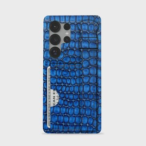 Może przedstawiać: Niebieskie etui na telefon z teksturowanym wzorem skóry krokodyla. Etui posiada pionową przegródkę na kartę z tyłu, zawierającą białą kartę z widocznym tekstem, oraz poziomy pasek w górnej części, służący do trzymania lub podparcia.