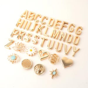 May include: Gold-colored metal alphabet letters and decorative charms. The letters are in a bold, block style. Charms include a lotus flower, daisy, heart, eye, and sunburst. The charms have turquoise and white accents.