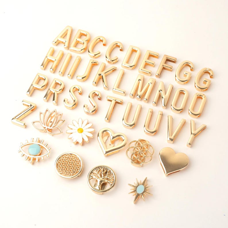 May include: Gold-colored metal alphabet letters and decorative charms. The letters are in a bold, block style. Charms include a lotus flower, daisy, heart, eye, and sunburst. The charms have turquoise and white accents.