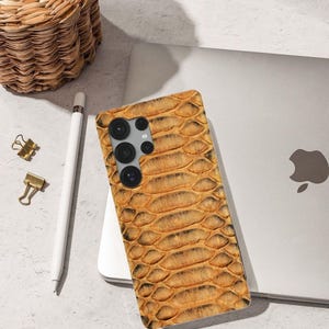 May include: A mobile phone case featuring a snakeskin design in brown and tan hues. The case rests on a white surface, alongside a laptop, a white stylus, and gold paper clips. A woven basket is visible in the background.