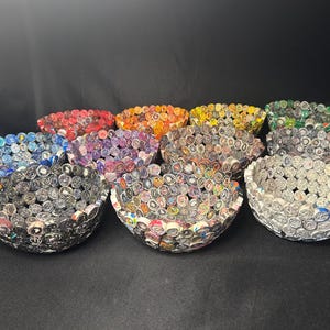 May include: A collection of handmade decorative bowls in various colors, including red, orange, yellow, green, blue, purple, and gray. Each bowl is crafted from rolled paper, creating a unique textured surface. The bowls are arranged on a dark surface.