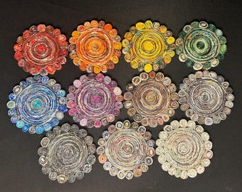 May include: A collection of eleven colorful, circular coasters. Each coaster is made from tightly rolled paper, creating a spiral design. The coasters feature a variety of colors, including red, orange, yellow, green, blue, purple, and neutral tones.