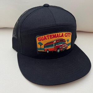 May include: Black trucker hat with a mesh back and a flat brim. The front panel features a patch with the words "GUATEMALA CITY" above an illustration of a red bus, trees, and a yellow background.