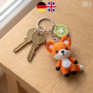 Peut inclure: Un porte-clés renard crocheté à la main, orange et blanc, avec des détails noirs et un charm en forme de fleur verte. Le porte-clés est attaché à un anneau argenté avec deux clés en laiton. Le renard mesure environ 7.6 cm de haut.