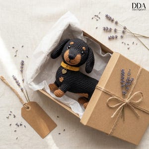 Peut inclure: Un chien teckel crocheté dans une boîte cadeau. Le chien noir et fauve porte un collier jaune avec un cœur. La boîte est marron et attachée avec de la ficelle, et contient des brins de lavande. L'image comprend une étiquette marron vierge et le texte "DDA Daisy DigitalMA".
