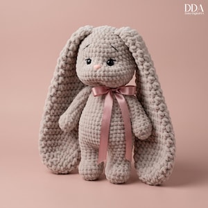 Peut inclure: Un lapin en crochet gris clair avec de longues oreilles et un nœud rose. Le jouet en peluche a des yeux noirs et un nez rose. Le lapin est posé sur un fond rose. Le logo "DDA Doris DigitalMA" est visible en haut à droite.