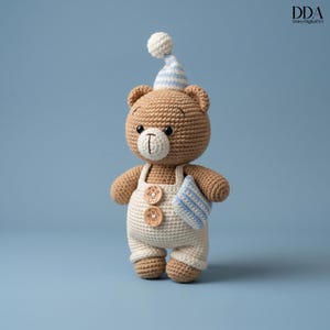 Peut inclure: Un ours en peluche crocheté portant un bonnet de nuit rayé bleu et blanc et une salopette crème avec des boutons en bois. L'ours tient un petit oreiller à rayures bleues et blanches. L'ours est brun et a des yeux noirs.