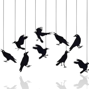 Könnte beinhalten: Eine Ansammlung schwarzer Krähensilhouetten, die an dünnen Schnüren vor einem weißen Hintergrund hängen. Die Krähen sind in verschiedenen Posen dargestellt, einige mit ausgebreiteten Flügeln, andere sitzen oder fliegen. Die Schatten der Krähen sind unten sichtbar.