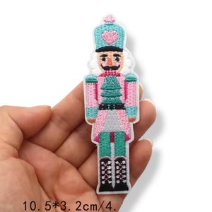 May include: Embroidered patch of a toy soldier nutcracker in pink, teal, and black. The patch features a detailed design with a teal hat, pink jacket, and black boots. The patch measures 4.1 inches in height and 1.3 inches in width.