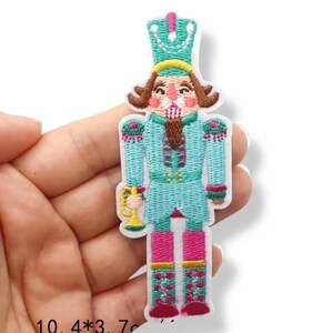 Może przedstawiać: Haftowana naszywka przedstawiająca żołnierza dziadka do orzechów w turkusowym mundurze z różowymi akcentami. Naszywka ma około 10,4 cm wysokości i 3,7 cm szerokości. Dziadek do orzechów trzyma złotą filiżankę i nosi turkusowy kapelusz.