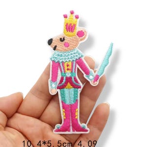 Może przedstawiać: Haftowana naszywka przedstawiająca postać myszy w różowo-turkusowym mundurze, w złotej koronie i z niebieskim mieczem. Naszywka ma 10,4 cm wysokości i 5,5 cm szerokości. Mysz ma różowy nos i uszy oraz czarne oczy.