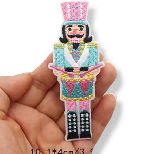 May include: Embroidered patch of a toy soldier nutcracker in pink, blue, and black. The nutcracker wears a pink hat, a blue jacket, and pink pants. The patch measures 10.1 x 4 cm.