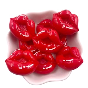 Op de afbeelding: Een verzameling glanzende, rood gevormde lippenbedels in een lichtroze, geschulpte schaal. De bedels hebben een glanzende afwerking met witte highlights, die op lippenstift lijken. De afbeelding is een close-up tegen een witte achtergrond.