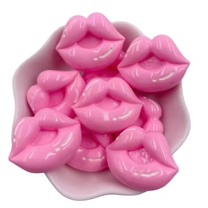 Op de afbeelding: Een verzameling glanzende, roze lipvormige versieringen in een lichtroze kom. De lippen hebben een glanzende afwerking met een subtiele witte highlight, waardoor een driedimensionaal effect ontstaat. De kom heeft een zachte, bloembladachtige vorm.