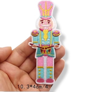 Może przedstawiać: Haftowana naszywka przedstawiająca żołnierza-dziadka do orzechów w kolorze różowym, niebieskim, żółtym i złotym. Naszywka ma około 10 cm wysokości i przedstawia szczegółowy wzór klasycznej postaci bożonarodzeniowej. Naszywka ma białą obwódkę.