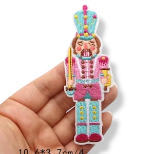 Może przedstawiać: Haftowana naszywka przedstawiająca żołnierza dziadka do orzechów w kolorze różowym, turkusowym i złotym. Naszywka przedstawia koronę, miecz i dekoracyjny mundur. Naszywka ma 10,6 cm wysokości i 3,7 cm szerokości.