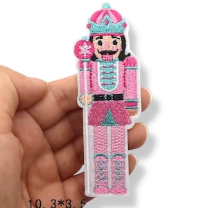 Może przedstawiać: Haftowana naszywka przedstawiająca różowego żołnierza-dziadka do orzechów z koroną i laską. Naszywka ma turkusowe i czarne akcenty, z tekstem "10.3*3.5" na dole. Naszywka ma około 10,3 cm wysokości i 3,5 cm szerokości.