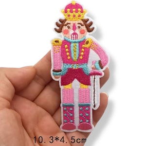 Op de afbeelding: Geborduurde patch van een notenkraker soldaat in roze, blauw en goud. De patch is 10,3 cm hoog en 4,5 cm breed. De notenkraker draagt een kroon en houdt een zwaard vast. De patch staat op een witte achtergrond.