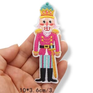 Może przedstawiać: Haftowana naszywka przedstawiająca żołnierza dziadka do orzechów w różowym mundurze ze złotymi akcentami, niebiesko-złotą koroną i mieczem. Naszywka ma około 10 cm wysokości i 3,6 cm szerokości.