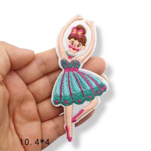 Könnte beinhalten: Gestickter Aufnäher einer Ballerina in einem türkis-rosa Kleid, Arme erhoben. Der Aufnäher ist ca. 10 cm x 10 cm groß. Die Ballerina hat braune Haare und eine rosa Schleife. Der Aufnäher befindet sich auf weißem Hintergrund.