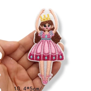 Może przedstawiać: Haftowana naszywka z baletnicą w różowej sukience ze złotą koroną. Naszywka ma około 10 cm wysokości i 5 cm szerokości. Baletnica ma brązowe włosy i różowe baletki.