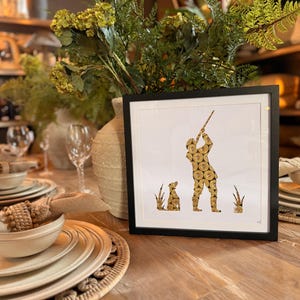 May include: A square framed print featuring a silhouette of a hunter aiming a shotgun with a dog sitting nearby, all composed of a mosaic pattern of gold and black shotgun shell casings against a stark white background within a black frame.