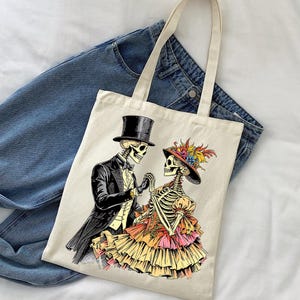 May include: Cream-colored tote bag featuring a Day of the Dead themed illustration. The graphic depicts a skeleton couple in formal attire, holding hands. The man wears a top hat and tuxedo, while the woman is in a ruffled dress and hat with feathers.