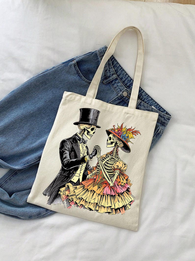 May include: Cream-colored tote bag featuring a Day of the Dead themed illustration. The graphic depicts a skeleton couple in formal attire, holding hands. The man wears a top hat and tuxedo, while the woman is in a ruffled dress and hat with feathers.
