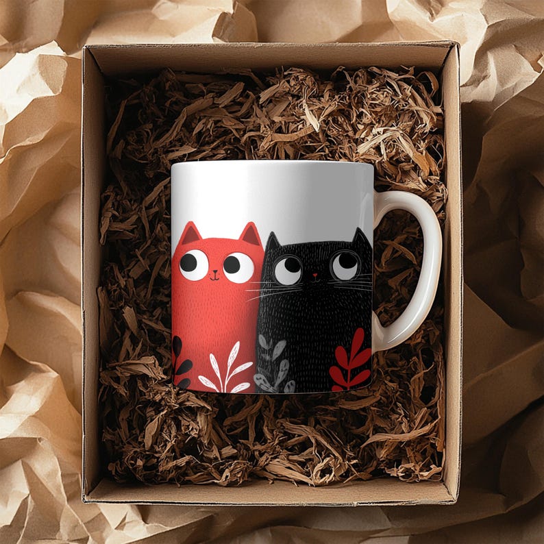 May include: White ceramic mug featuring a red and black cat illustration. The red cat has a smiling face and the black cat has a textured design. The mug is inside a cardboard gift box with shredded paper.