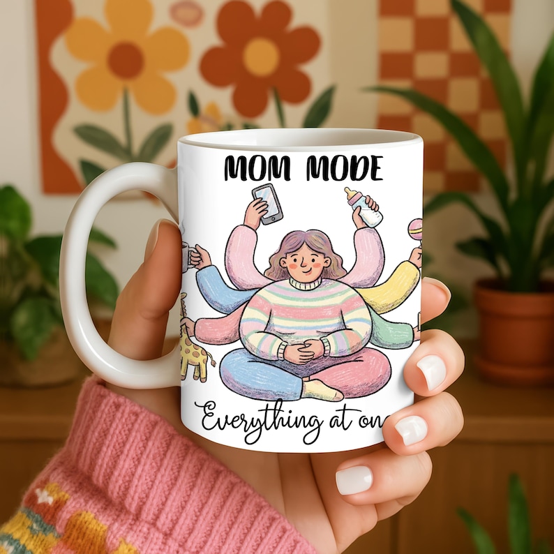 K&ouml;nnte beinhalten: Wei&szlig;e Keramik-Tasse mit den schwarzen Worten "MOM MODE". Die Tasse zeigt eine Cartoon-Illustration einer Person mit mehreren Armen, die verschiedene Gegenst&auml;nde h&auml;lt. Der Text "Everything at on" ist ebenfalls sichtbar.