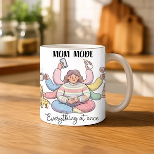K&ouml;nnte beinhalten: Wei&szlig;e Keramik-Tasse mit den Worten "MOM MODE" und "Everything at once" aufgedruckt. Die Tasse zeigt eine Cartoon-Illustration einer Frau mit mehreren Armen, die verschiedene Gegenst&auml;nde h&auml;lt, darunter ein Telefon, eine Babyflasche und eine Kaffeetasse.