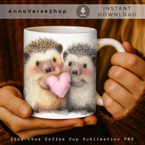 May include: White ceramic coffee mug featuring two adorable hedgehogs holding a pink heart. The mug has a white handle and the text "AnnaVerseShop" and "INSTANT DOWNLOAD" are printed on the mug.