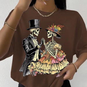 May include: Brown t-shirt featuring a skeletal couple in formal attire, holding hands. The male skeleton wears a top hat and tuxedo, while the female skeleton dons a hat with floral accents and a layered dress. The design is a Day of the Dead theme.
