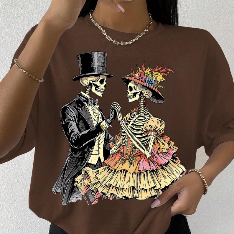 May include: Brown t-shirt featuring a skeletal couple in formal attire, holding hands. The male skeleton wears a top hat and tuxedo, while the female skeleton dons a hat with floral accents and a layered dress. The design is a Day of the Dead theme.