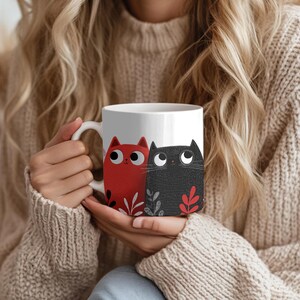 May include: White ceramic mug featuring a red and a black cat illustration. The cats have large eyes and are decorated with leaf accents. The mug has a curved handle and is held by a person wearing a beige sweater.