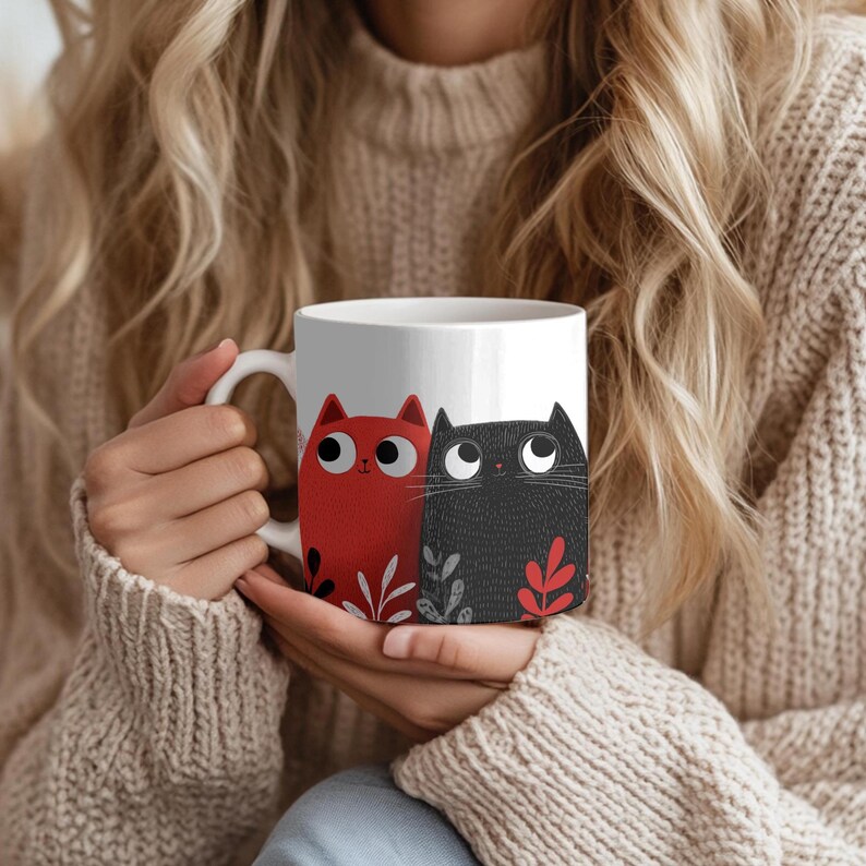 May include: White ceramic mug featuring a red and a black cat illustration. The cats have large eyes and are decorated with leaf accents. The mug has a curved handle and is held by a person wearing a beige sweater.