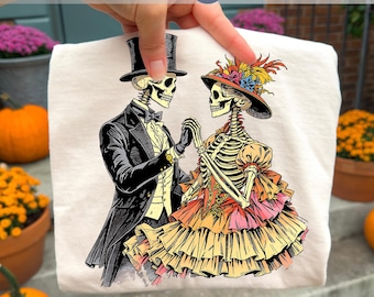 May include: A white t-shirt featuring a vintage illustration of two skeletons dressed in formal attire, holding hands. The male skeleton wears a top hat and tuxedo, while the female skeleton is in a dress and hat. The text "HIGH QUALITY PNG" is at the bottom.