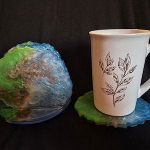 Puede incluir: Una taza de cerámica blanca con un diseño de hojas negras descansa sobre un posavasos de resina azul y verde. Hay otro posavasos a juego al lado. Los posavasos tienen un aspecto veteado.