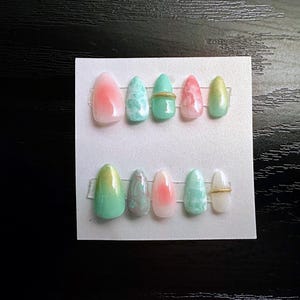 May include: A set of ten artificial nails in various colors, including pink, green, and white. Some nails feature a marbled design, while others have a gold band. The nails are arranged on a white card.