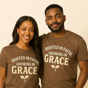 Puede incluir: Dos personas con camisetas marrones con el texto "ROOTED IN FAITH, GROWING IN GRACE" en blanco. Las camisetas también presentan un pequeño gráfico de hojas. Llevan vaqueros azules.