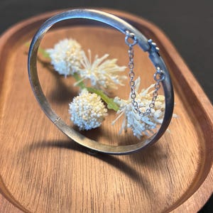 May include: A silver-toned hinged bangle bracelet with a delicate chain detail. The bracelet is displayed on a wooden tray with white, spherical flowers. The bracelet's design is simple and elegant, suitable for everyday wear.