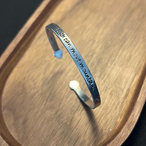 Puede incluir: Una pulsera de plata con la inscripción grabada "Faith can move mountains". La pulsera abierta está colocada sobre una bandeja ovalada de madera. La pulsera tiene un detalle en forma de corazón.
