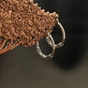 Pode incluir: Brincos de argola prateados com um design decorativo preto e prateado. Os brincos são exibidos em um porta-joias marrom em forma de flor contra um fundo escuro. Os brincos têm formato circular.