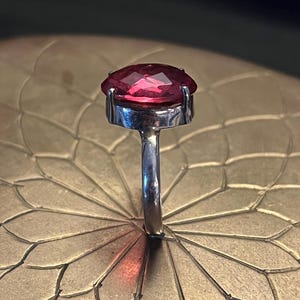 Pode incluir: Um anel de prata com uma grande pedra preciosa vermelha escura facetada em forma oval. A pedra preciosa est&aacute; engastada numa moldura simples e cl&aacute;ssica. O anel &eacute; exibido numa superf&iacute;cie dourada texturizada.