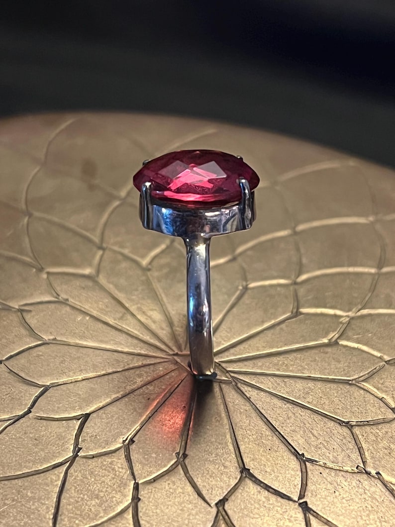 Pode incluir: Um anel de prata com uma grande pedra preciosa vermelha escura facetada em forma oval. A pedra preciosa est&aacute; engastada numa moldura simples e cl&aacute;ssica. O anel &eacute; exibido numa superf&iacute;cie dourada texturizada.